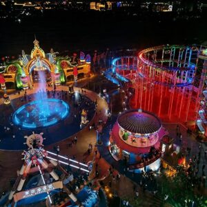 Aerial night view of an amusement park featuring an illuminated pirate ship ride, carousel, roller coaster, and fountain plaza