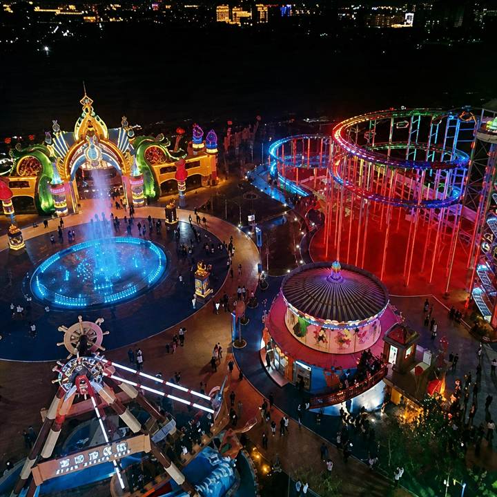 Aerial night view of an amusement park featuring an illuminated pirate ship ride, carousel, roller coaster, and fountain plaza
