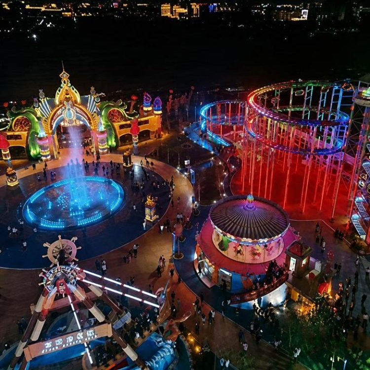 Aerial night view of an amusement park featuring an illuminated pirate ship ride, carousel, roller coaster, and fountain plaza