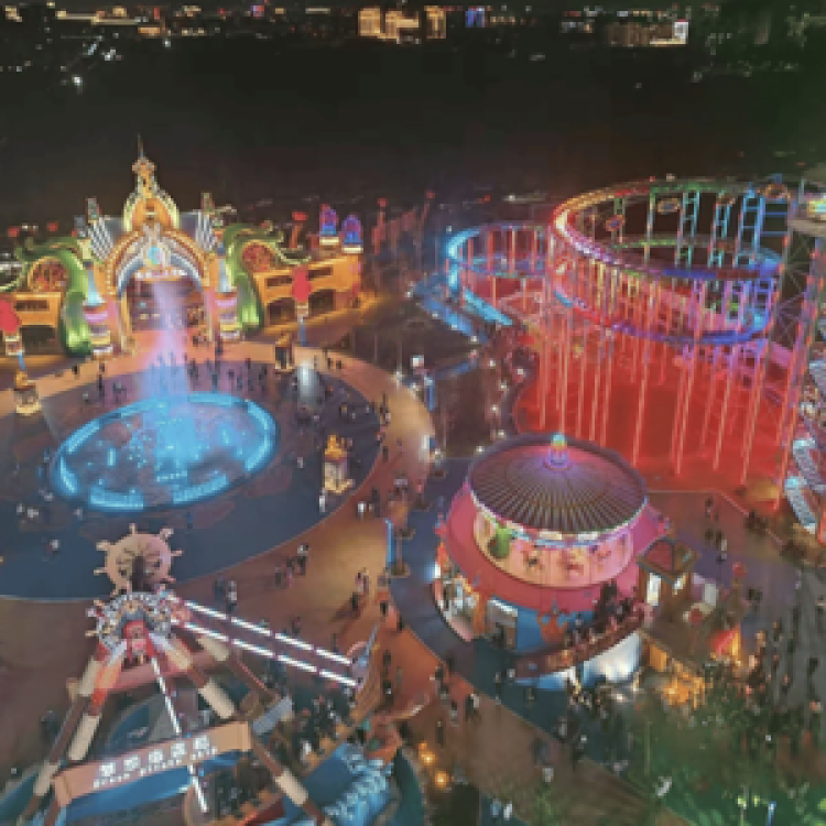 Aerial night view of an amusement park featuring an illuminated pirate ship ride, carousel, roller coaster, and fountain plaza